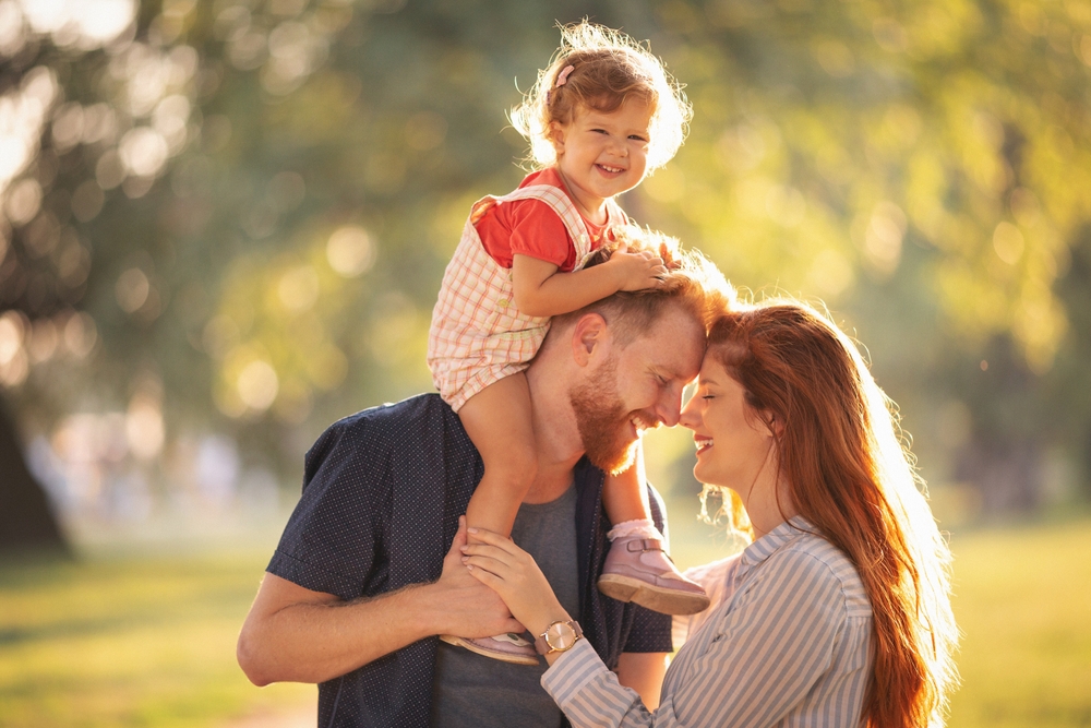 Parents smiling together outdoors with their daughter on her father’s shoulders
